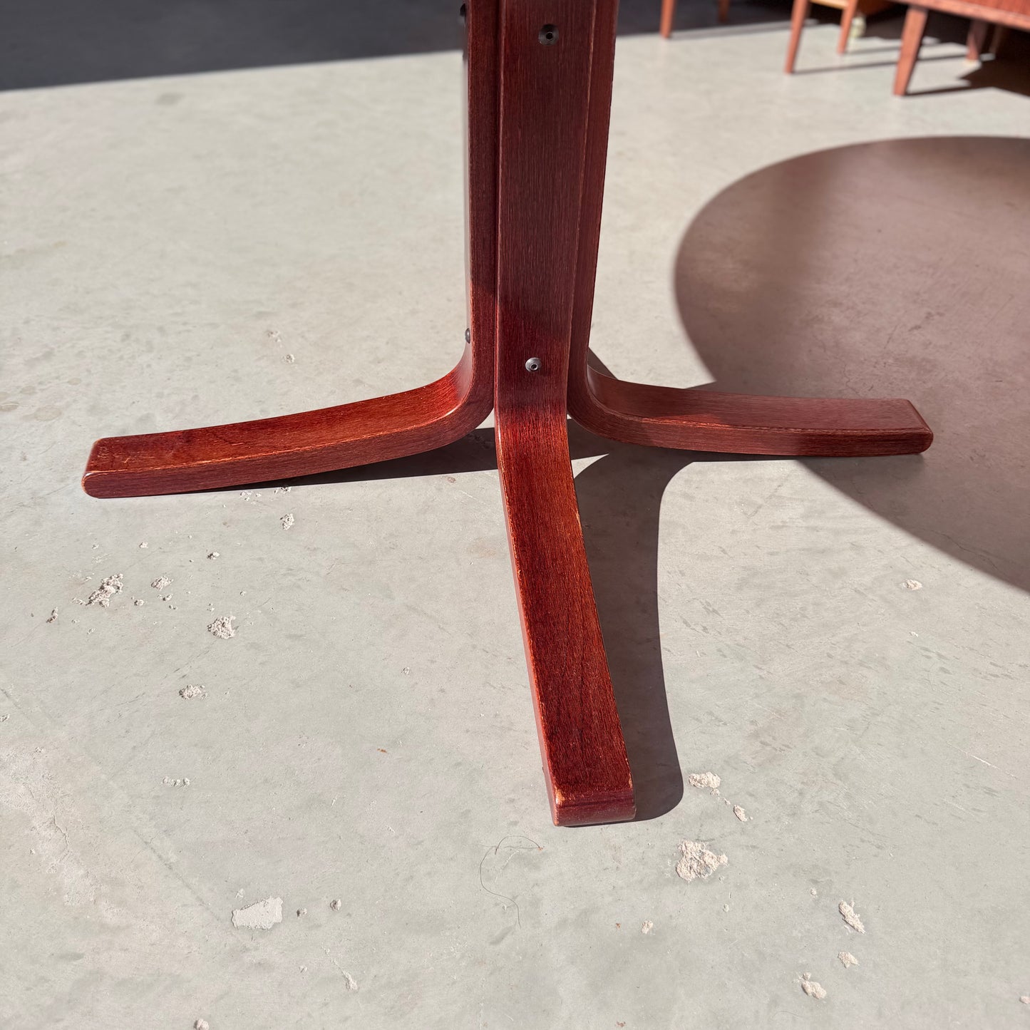 Close-up of the solid wood base on the Joe Rufenacht Designer Round Dining Table – Rare Mid-Century Gem, showing its three-legged design on a sunlit light gray floor with a round shadow and worn spots adding unique character.