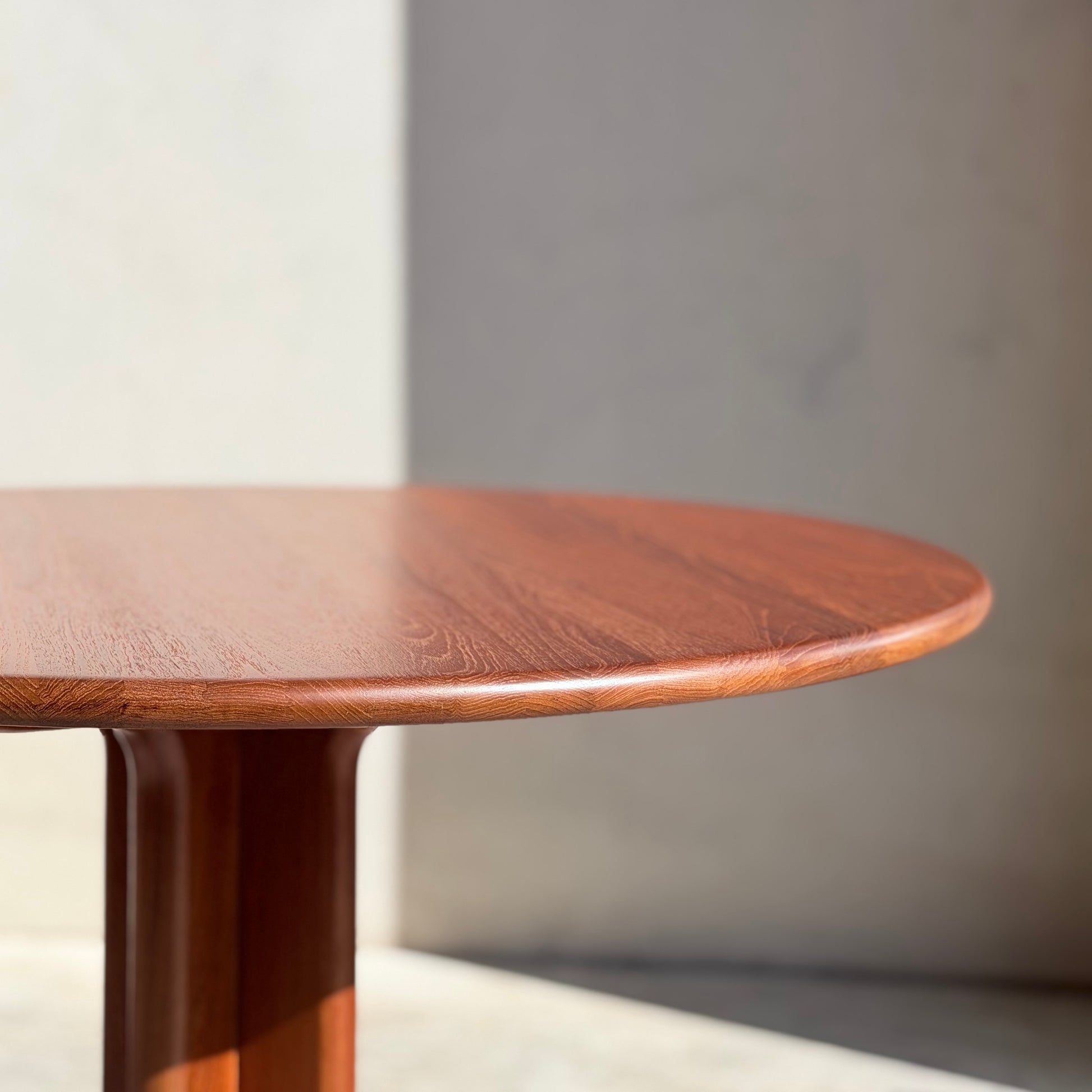 A close-up of the Fully Restored Solid Teak Danish Dining Table (Rediscover), featuring a polished round surface and central leg, set against a plain light wall with soft natural light and shadows.