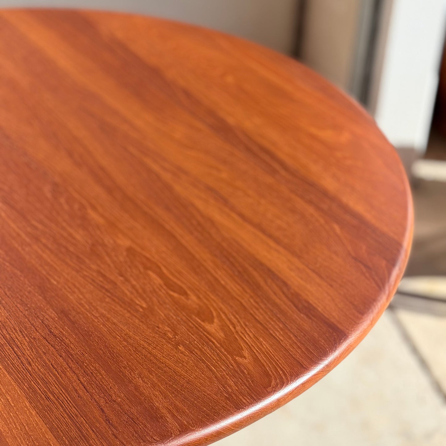 A close-up of the Fully Restored Solid Teak Danish Dining Table (Rediscover) showcases its smooth, polished surface and striking wood grain illuminated by natural light near a doorway.
