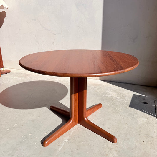 A Fully Restored Solid Teak Danish Dining Table (Rediscover) with a central pedestal base and rich brown finish stands on a smooth concrete floor, sunlight casting shadows against a plain, light-colored wall.