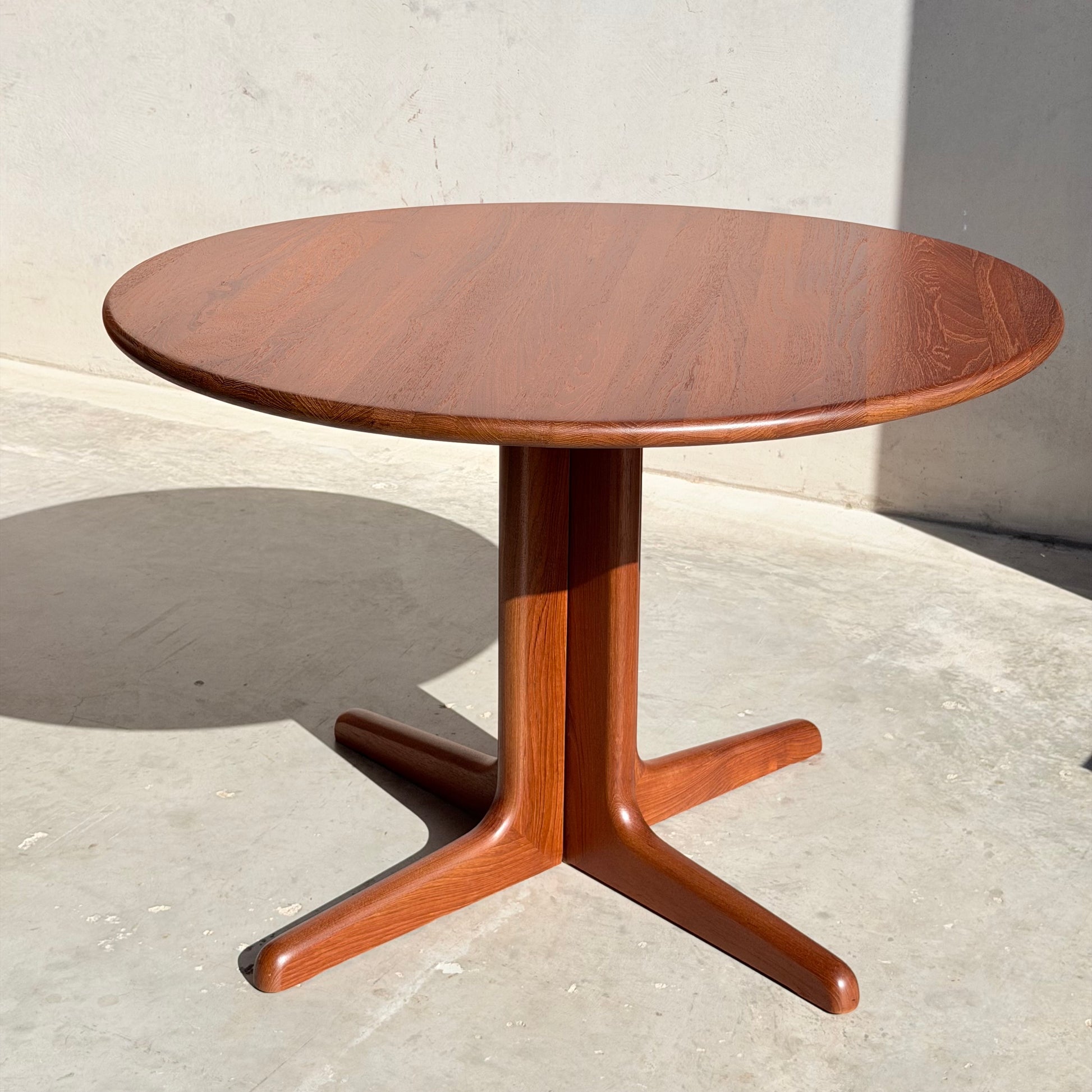 A Fully Restored Solid Teak Danish Dining Table (Rediscover) with a sleek, three-legged pedestal base stands on a concrete floor by a light wall, sunlight accentuating its mid-century modern charm and smooth vintage finish.