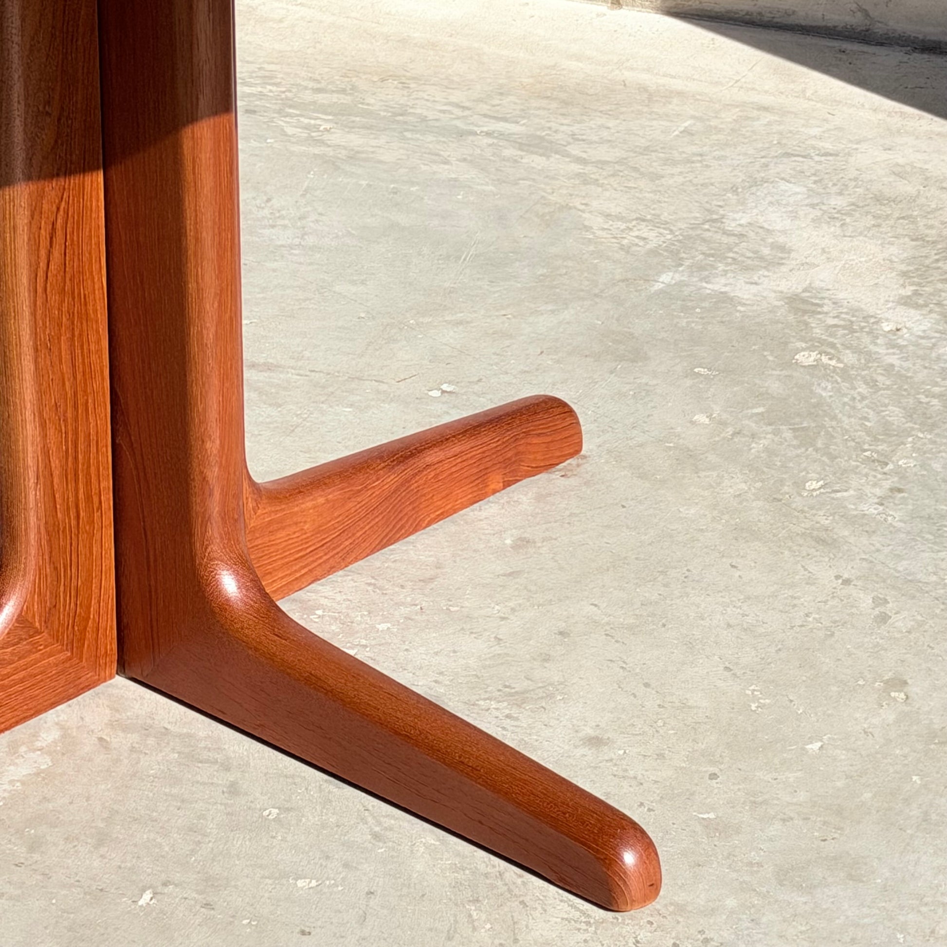 Two solid teak legs of the Fully Restored Solid Teak Danish Dining Table (Rediscover) cast shadows on a smooth, light concrete floor as sunlight highlights their rich reddish-brown finish and wood grain.
