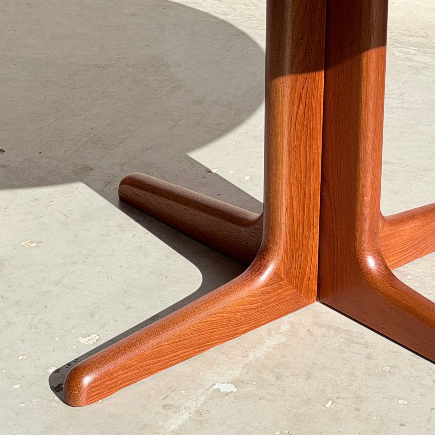 Close-up of the wooden base on the Fully Restored Solid Teak Danish Dining Table (Rediscover), with two V-shaped legs casting shadows on a light floor. The solid teak wood features a warm, reddish-brown hue.