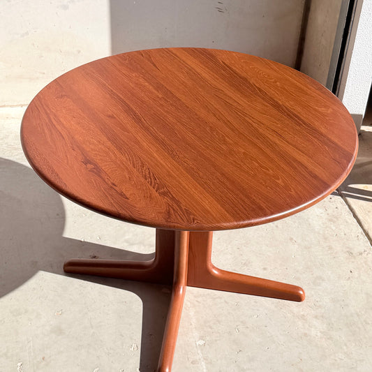 The Fully Restored Solid Teak Danish Dining Table (Rediscover) features a smooth finish and single pedestal base, standing on a light concrete floor and beautifully illuminated by bright sunlight.