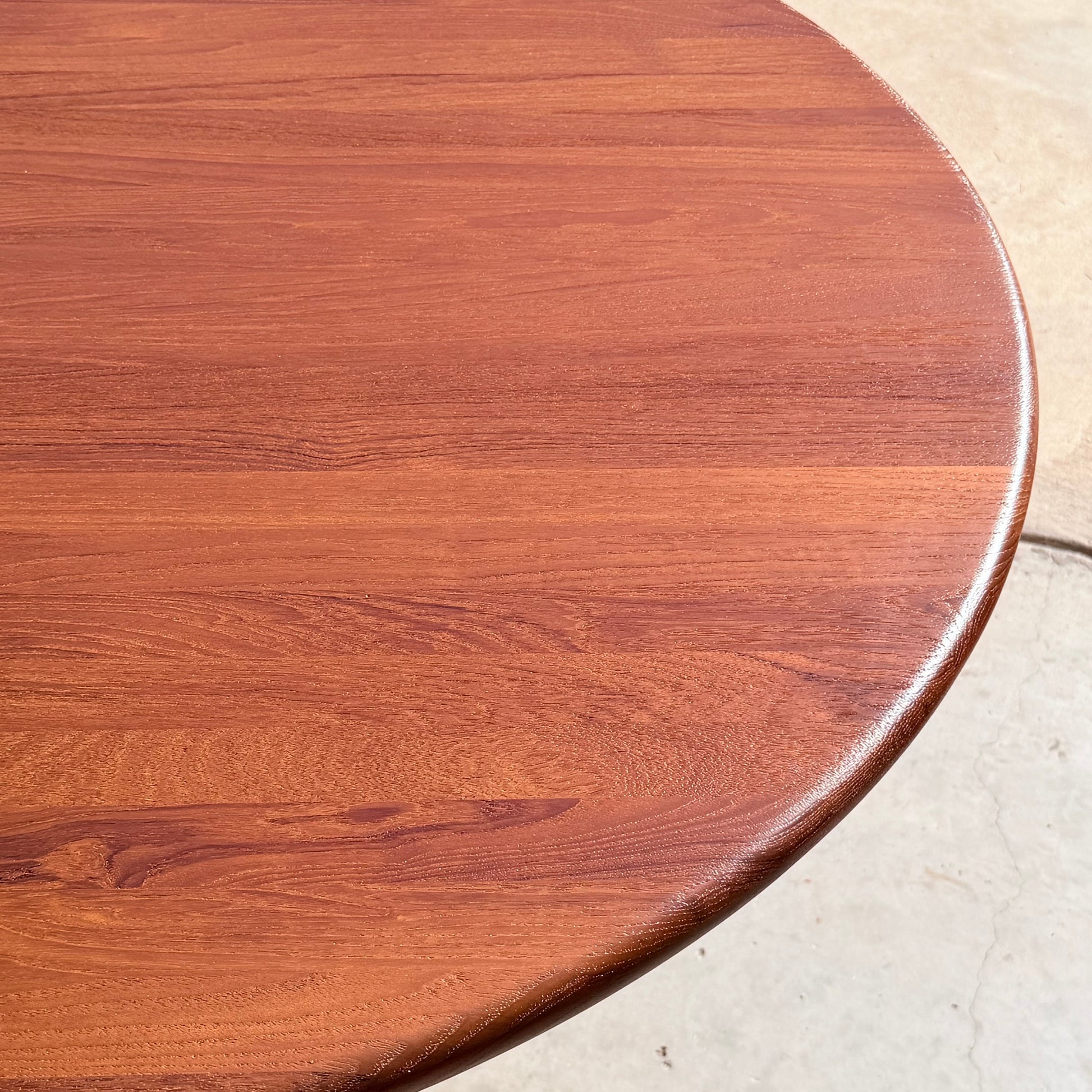 A close-up of the Fully Restored Solid Teak Danish Dining Table (Rediscover), featuring a smooth, polished round surface with visible wood grain patterns, set on a light-colored floor.