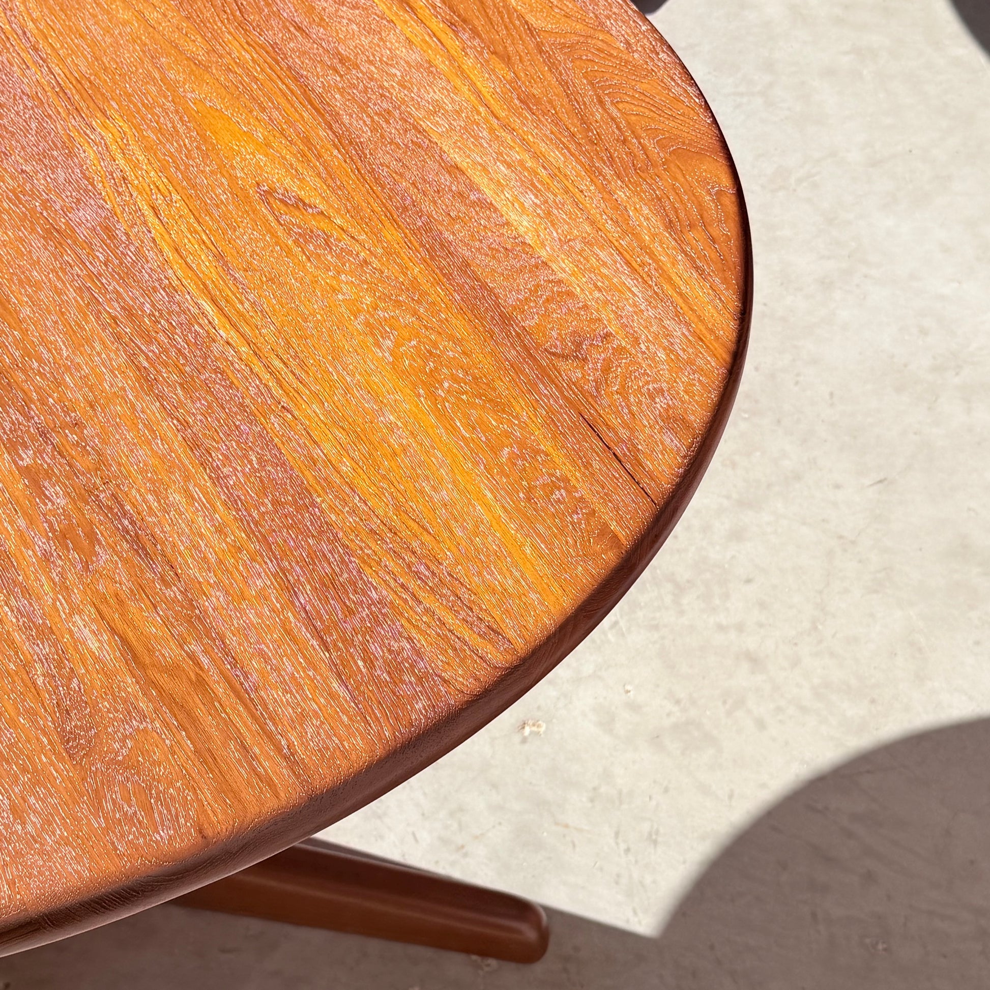 A close-up of the Fully Restored Solid Teak Danish Dining Table (Rediscover) shows its smooth, polished round surface with visible wood grain, casting a soft shadow on a light floor.