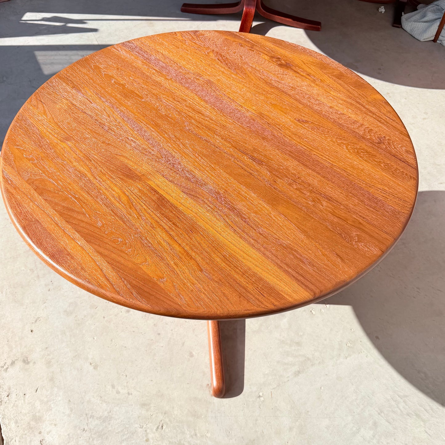 A Fully Restored Solid Teak Danish Dining Table (Rediscover), with a rich reddish-brown finish and visible wood grain, sits on a light concrete floor, sunlight highlighting its Danish modern design.