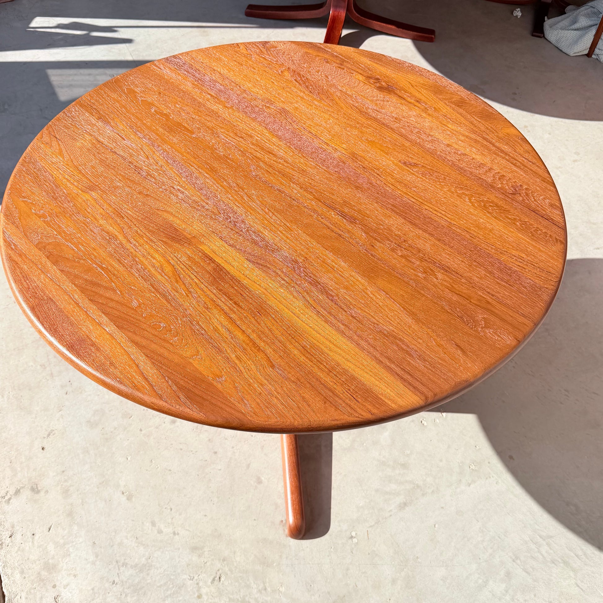 A Fully Restored Solid Teak Danish Dining Table (Rediscover), with a rich reddish-brown finish and visible wood grain, sits on a light concrete floor, sunlight highlighting its Danish modern design.