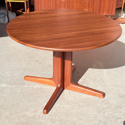 The Fully Restored Solid Teak Danish Dining Table (Rediscover) features a vintage round design with a smooth finish and central pedestal base splitting into three curved legs, set on a light-colored concrete floor.