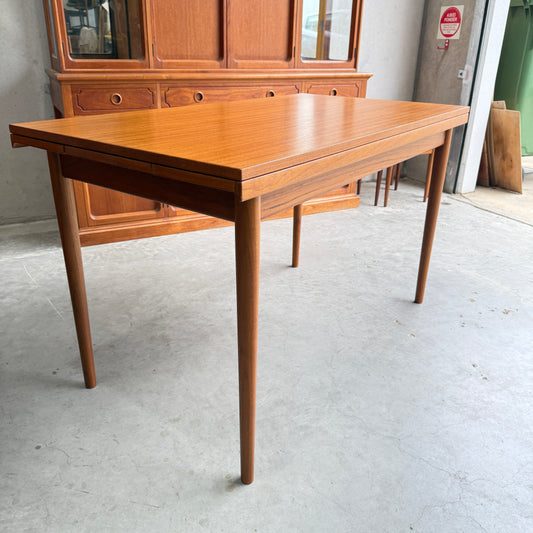 A Fully Restored Parker 95 Teak Nordic Extension Table (Rediscover) with tapered legs sits on a concrete floor, with a matching wooden hutch in the background against the wall.