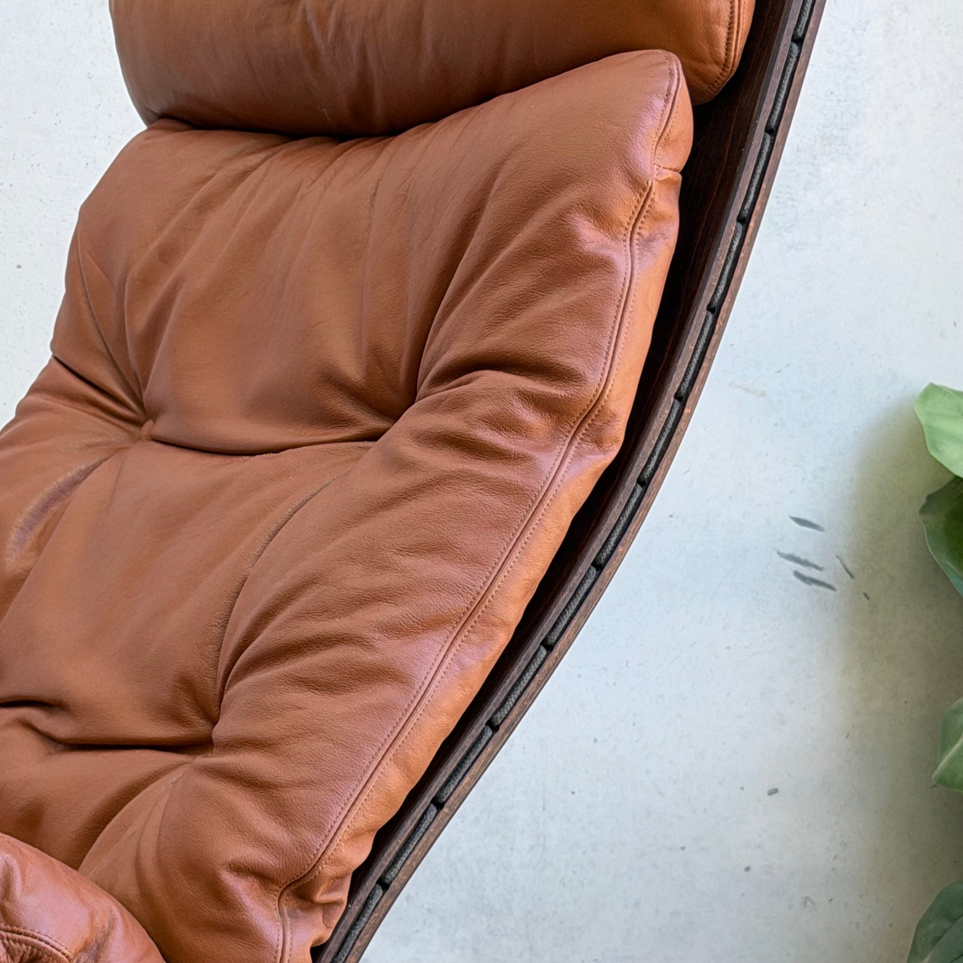 A close-up of the Signature Vintage Ingmar Relling Siesta Set—two brown leather lounge chairs with tufted cushions—inspired by mid-century Scandinavian design, beside a light wall and a green plant visible on the right.