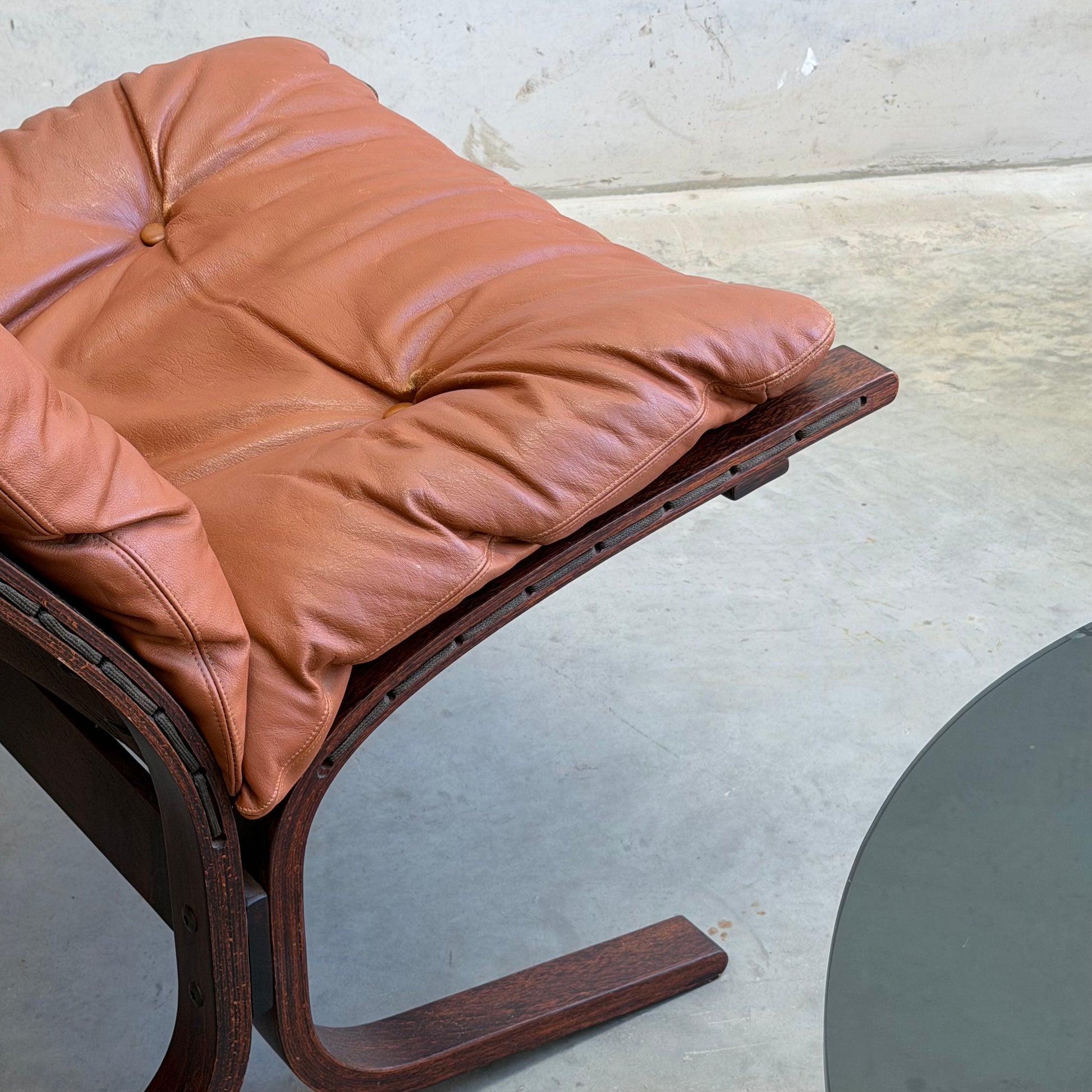 A close-up of the Signature Vintage Ingmar Relling Siesta Set—two lounge chairs with dark curved wood and tufted brown leather, paired with a rare round glass coffee table—sits on a concrete floor.