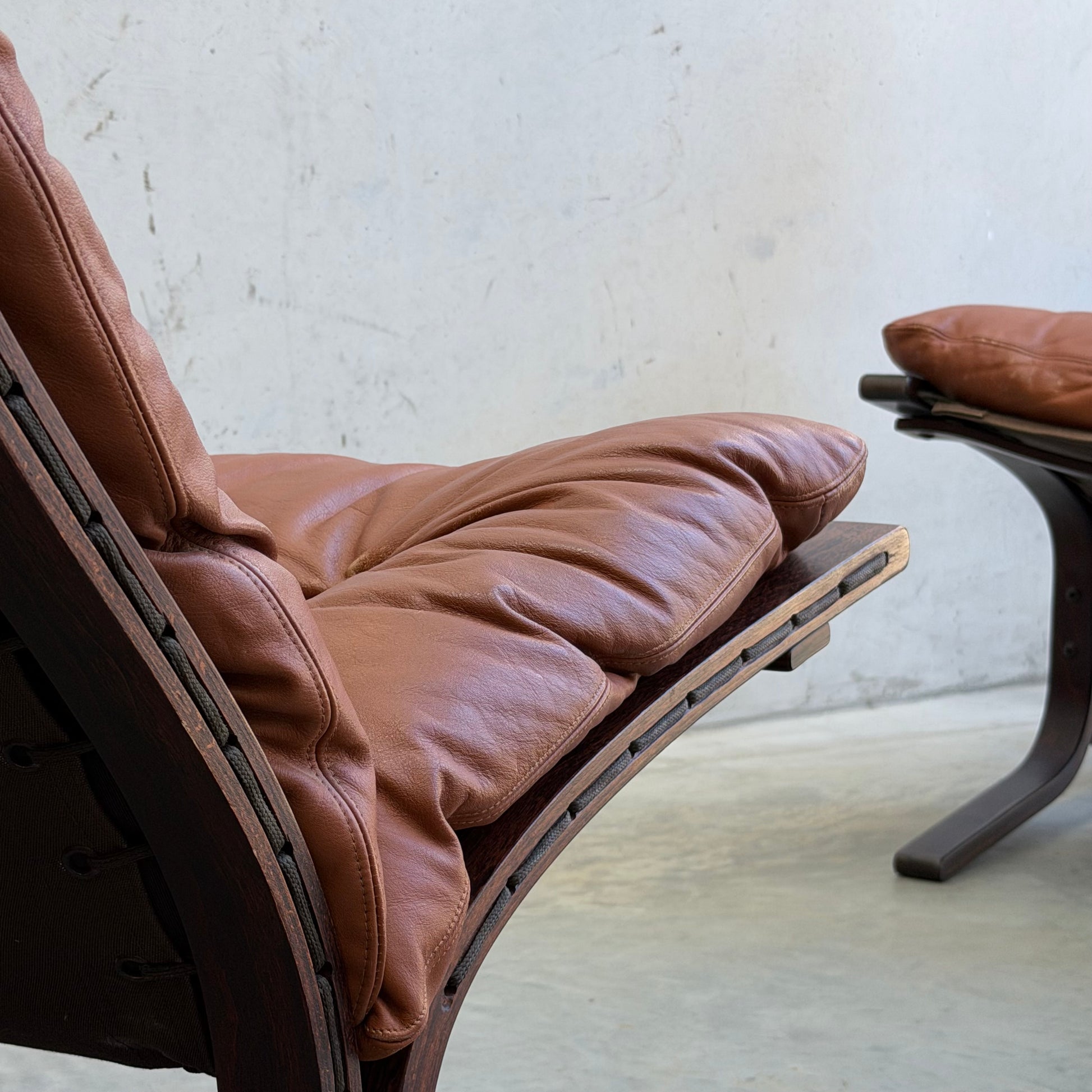 Close-up of the Signature Vintage Ingmar Relling Siesta Set—two lounge chairs with plush brown leather cushions and dark curved wood, plus a rare coffee table—a classic example of Scandinavian mid-century furniture against a plain wall on concrete.