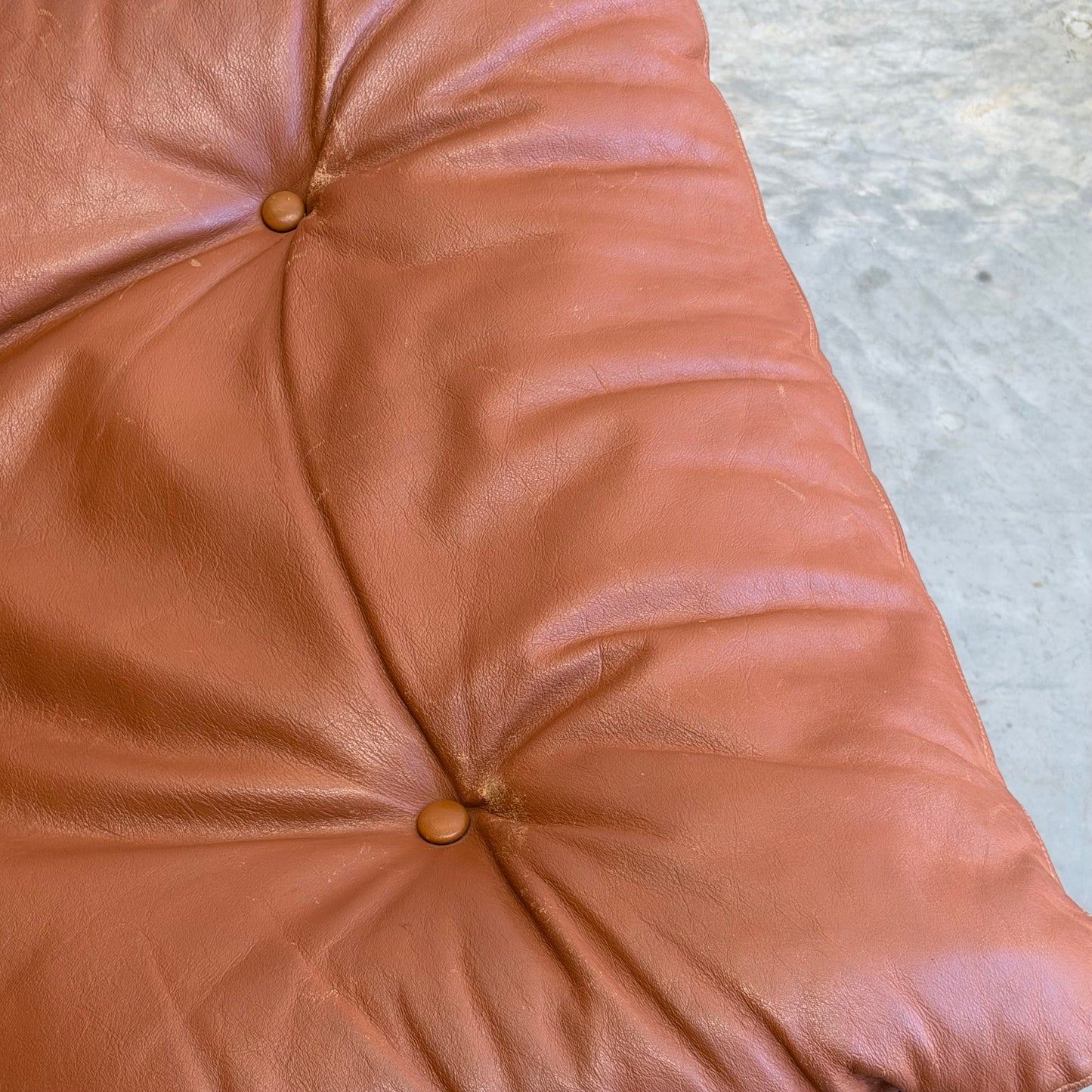 Close-up of a tufted brown leather cushion with two buttons from the Signature Vintage Ingmar Relling Siesta Set—Two Lounge Chairs & Rare Coffee Table—resting on a gray, textured concrete surface.