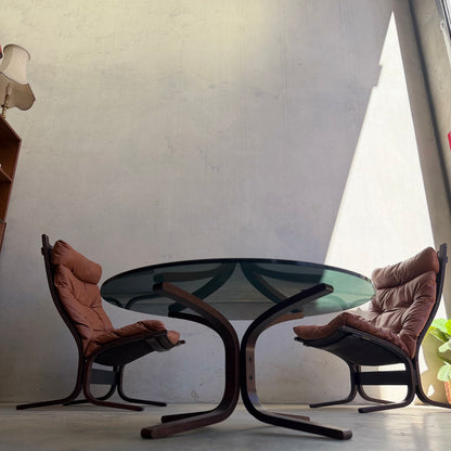 A Signature Vintage Ingmar Relling Siesta Set—two cushioned brown lounge chairs and a rare coffee table—stands in a minimalist room with tall concrete walls, bright sunlight, and glimpses of a lamp and bookshelf on the left.