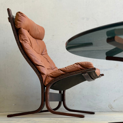 The Signature Vintage Ingmar Relling Siesta Set features two lounge chairs with brown tufted leather cushions and curved dark wood frames, paired with a rare round glass coffee table, embodying Scandinavian mid-century style.