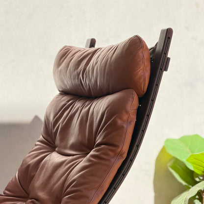 A close-up of the Signature Vintage Ingmar Relling Siesta Set—two modern brown leather lounge chairs with plush cushions and a rare coffee table, set against a light background, channels Scandinavian mid-century style. A green leafy plant appears partially on the right.