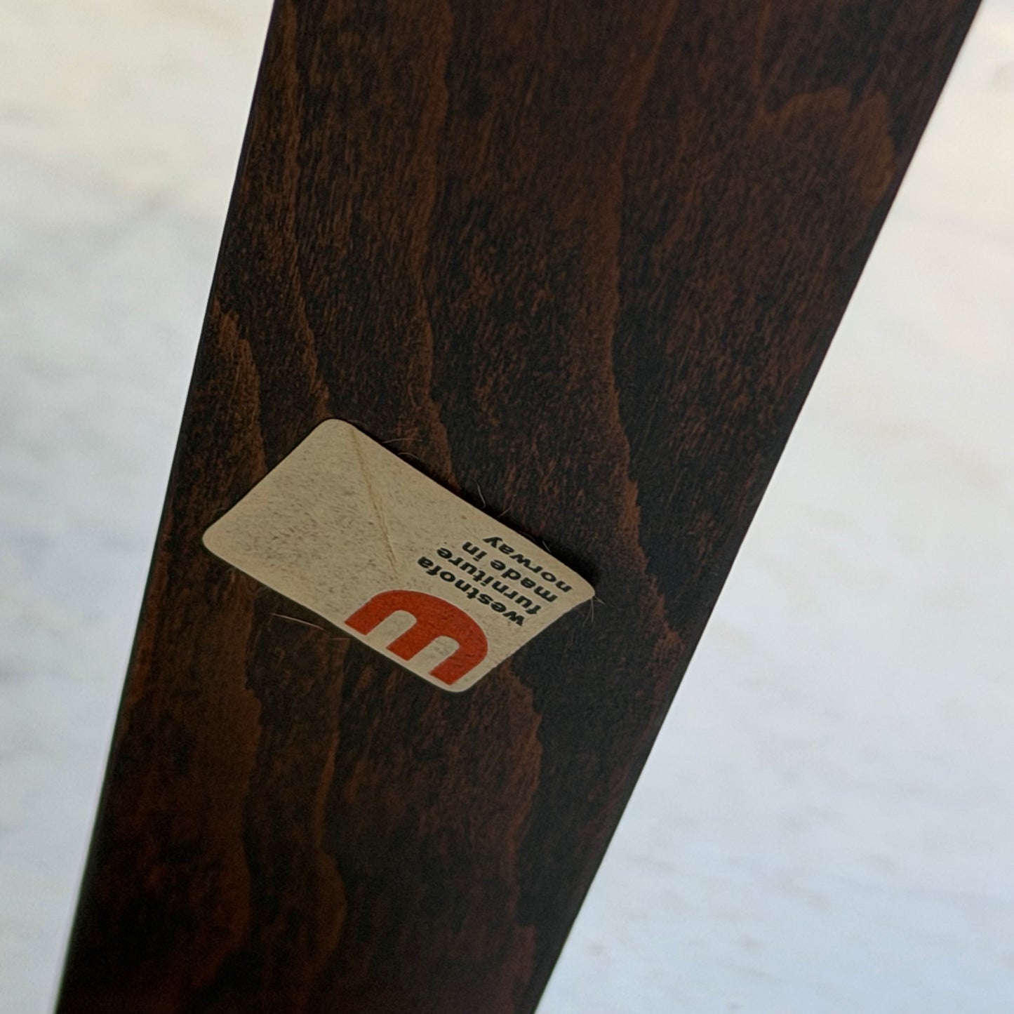 A close-up of a wooden furniture leg from the Signature Vintage Ingmar Relling Siesta Set – Two Lounge Chairs & Rare Coffee Table, showing an upside-down rectangular sticker with a red m logo and some partially visible text; background blurred.