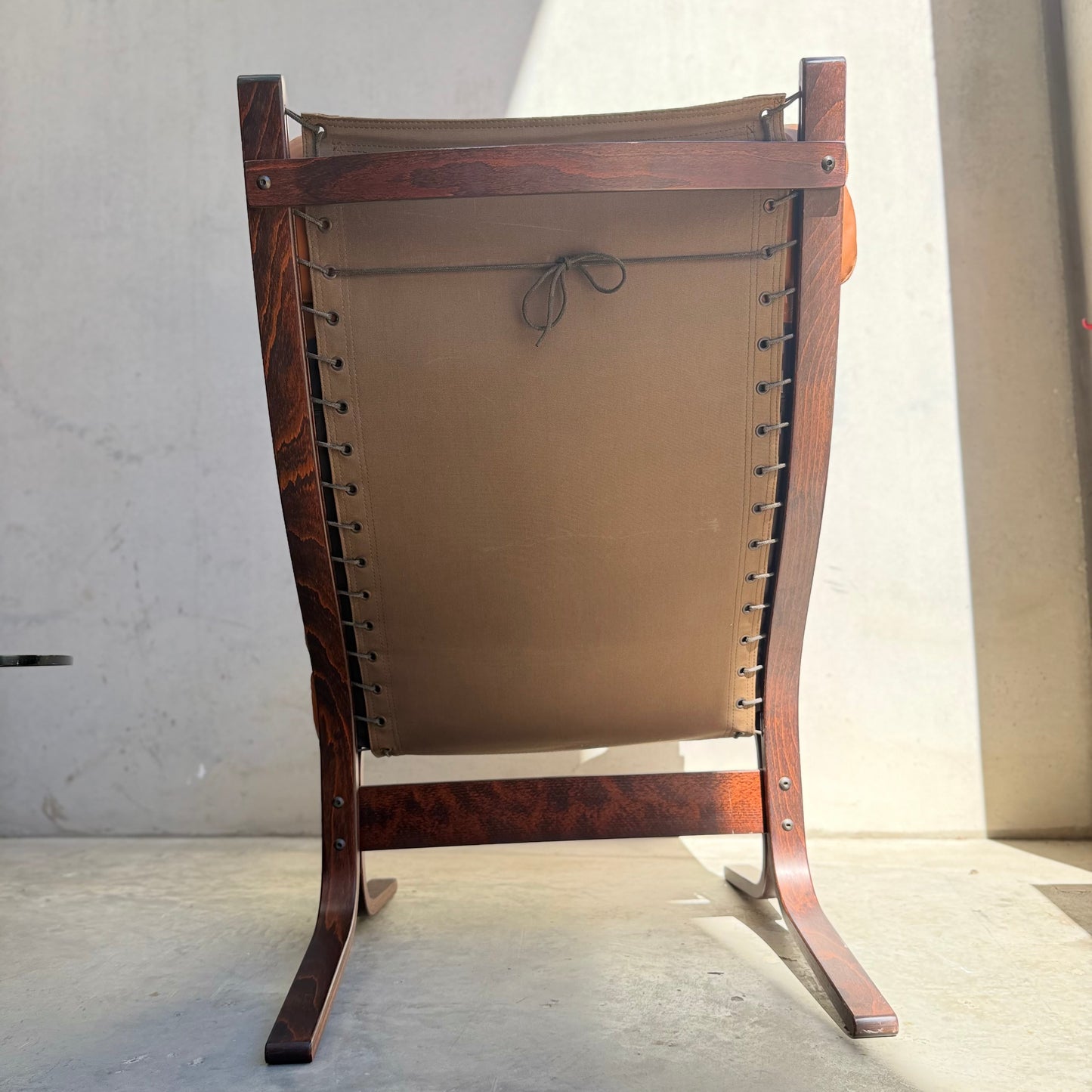 Viewed from behind, sunlight highlights the curved legs of the Signature Vintage Ingmar Relling Siesta Set—two lounge chairs with wooden frames and brown canvas backs, iconic of Scandinavian mid-century design.