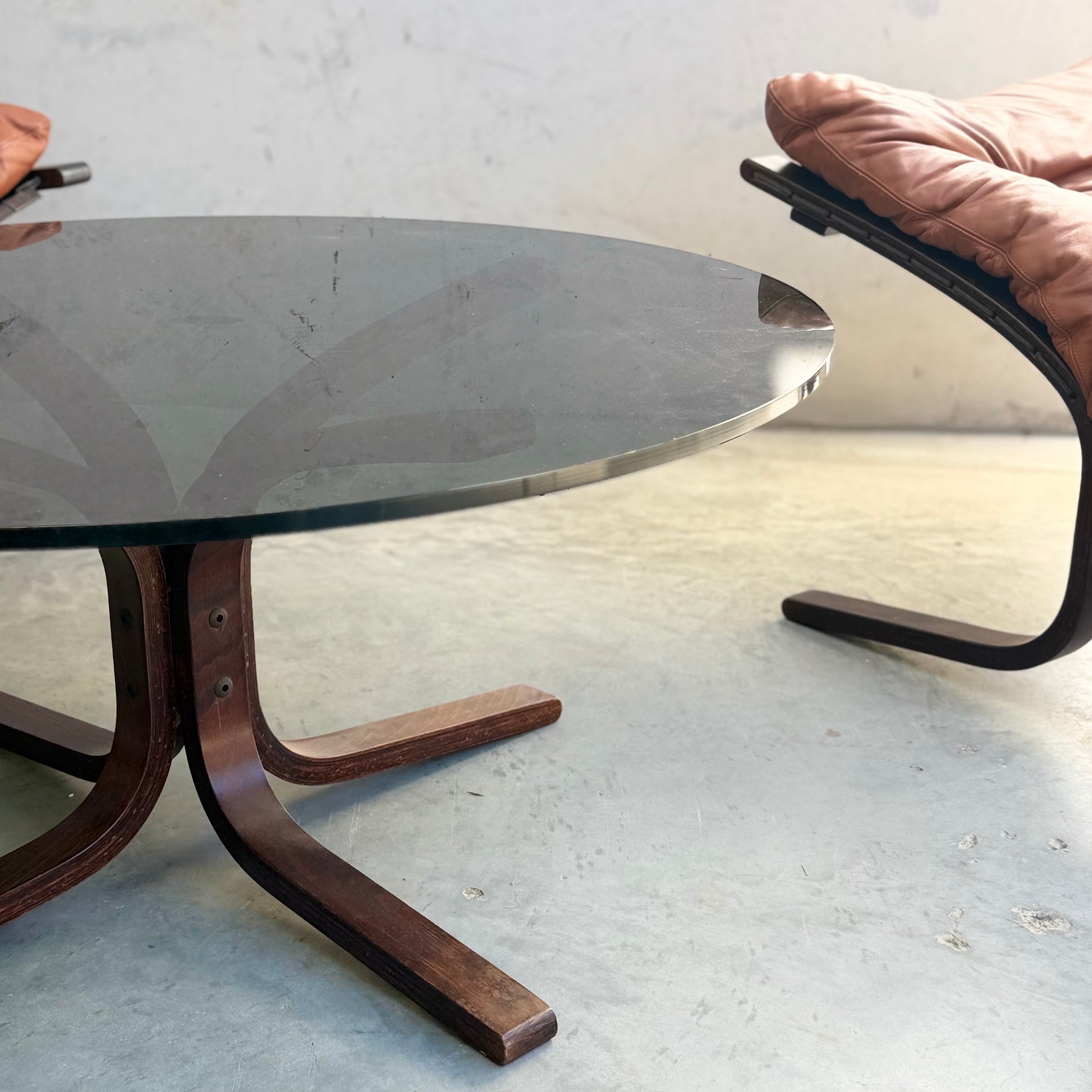 A Signature Vintage Ingmar Relling Siesta Set—two lounge chairs with wooden frames and tan cushions, plus a rare modern glass coffee table—evokes classic Scandinavian mid-century style on a light concrete floor.
