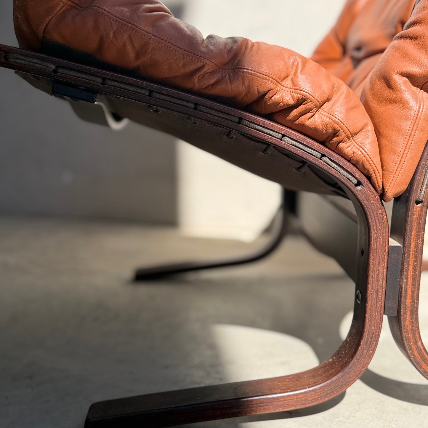 Close-up side view of the Signature Vintage Ingmar Relling Siesta Set lounge chair, featuring a curved dark wood frame and tufted brown leather cushions, iconic Scandinavian mid-century design, shown on a sunlit concrete floor.