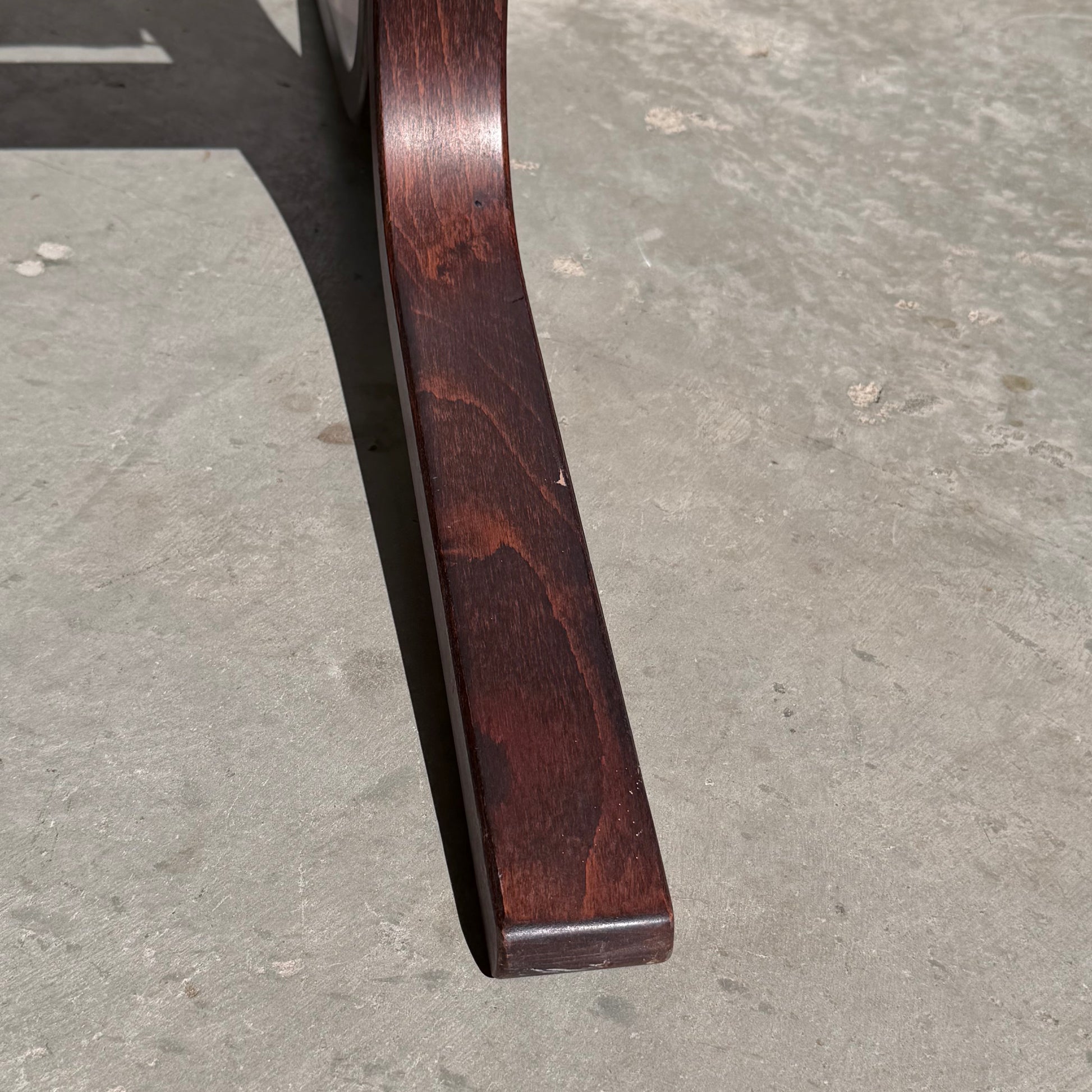 A close-up of the curved wooden leg from the Signature Vintage Ingmar Relling Siesta Set lounge chair, echoing Scandinavian mid-century style, with polished wood grain and subtle imperfections casting a shadow on a gray concrete floor.