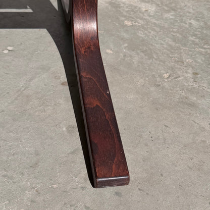 A close-up of the curved wooden leg from the Signature Vintage Ingmar Relling Siesta Set lounge chair, echoing Scandinavian mid-century style, with polished wood grain and subtle imperfections casting a shadow on a gray concrete floor.
