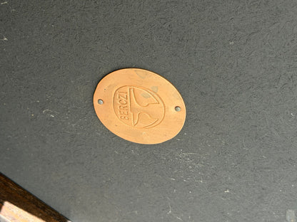 A circular hand-hammered copper plaque reading BERGZI and featuring a simple logo is affixed with two screws to the dark, textured surface of the Vintage Louis Berczi Hammered Copper Round Coffee Table – Rare Australian Collectible (Revive).