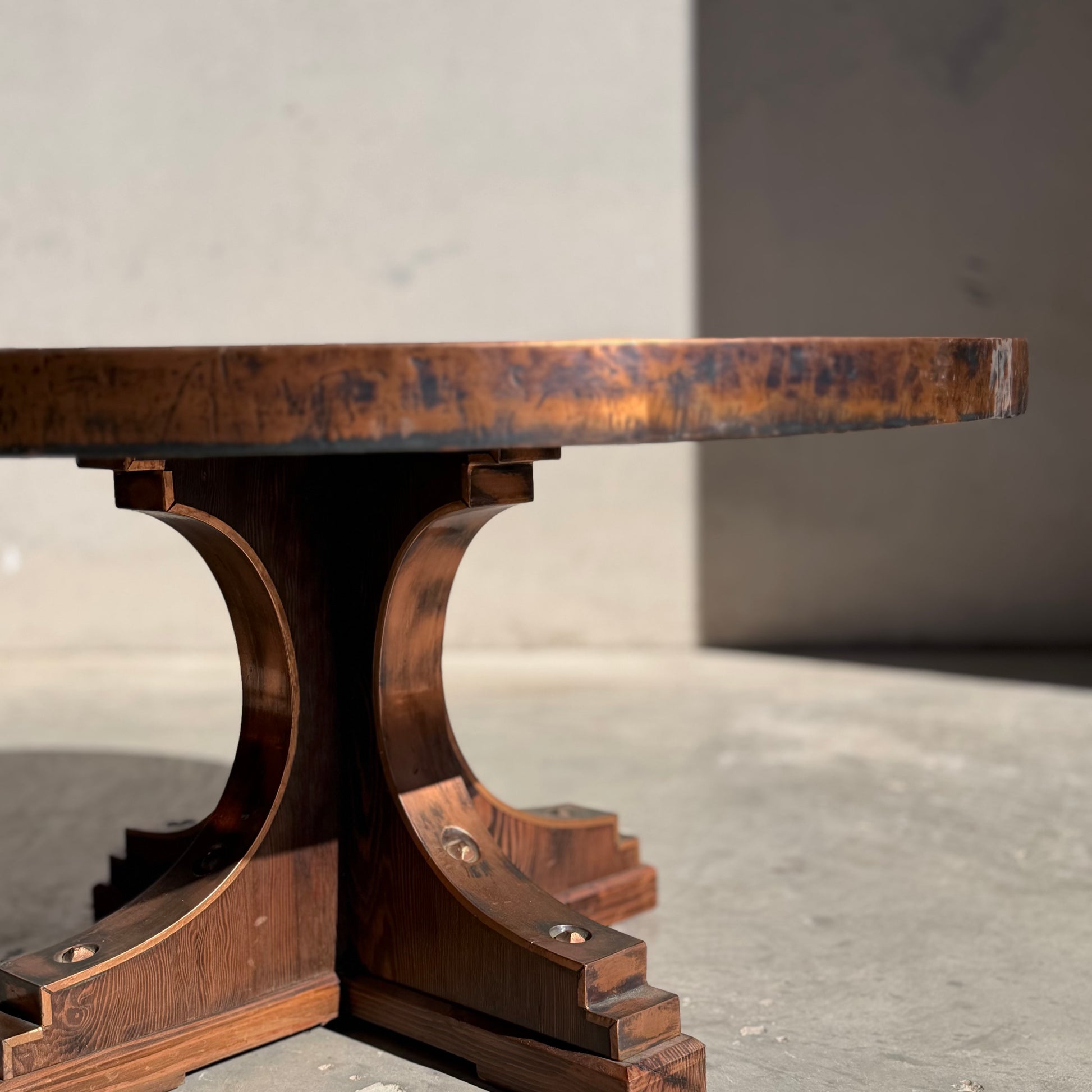 A close-up, low-angle view of the Vintage Louis Berczi Hammered Copper Round Coffee Table, a rare Australian collectible, featuring a thick wooden top and ornate curved pedestal base casting shadows on a plain, light wall.
