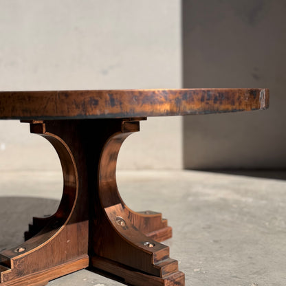 A close-up, low-angle view of the Vintage Louis Berczi Hammered Copper Round Coffee Table, a rare Australian collectible, featuring a thick wooden top and ornate curved pedestal base casting shadows on a plain, light wall.