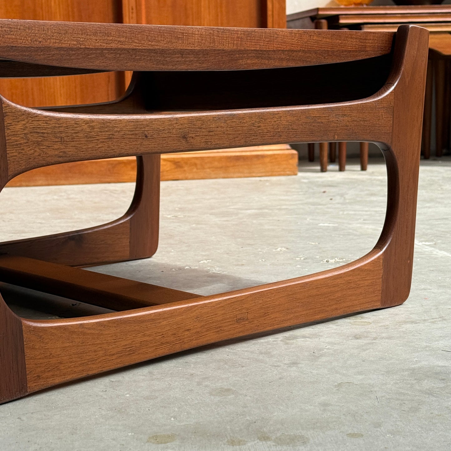 Close-up of an ultra rare Gerald Easden coffee table in Blackwood with a sleigh leg base (Rediscover), mid-century modern style, featuring curved edges and smooth finish on a concrete floor; another wooden piece is partially visible in the background.