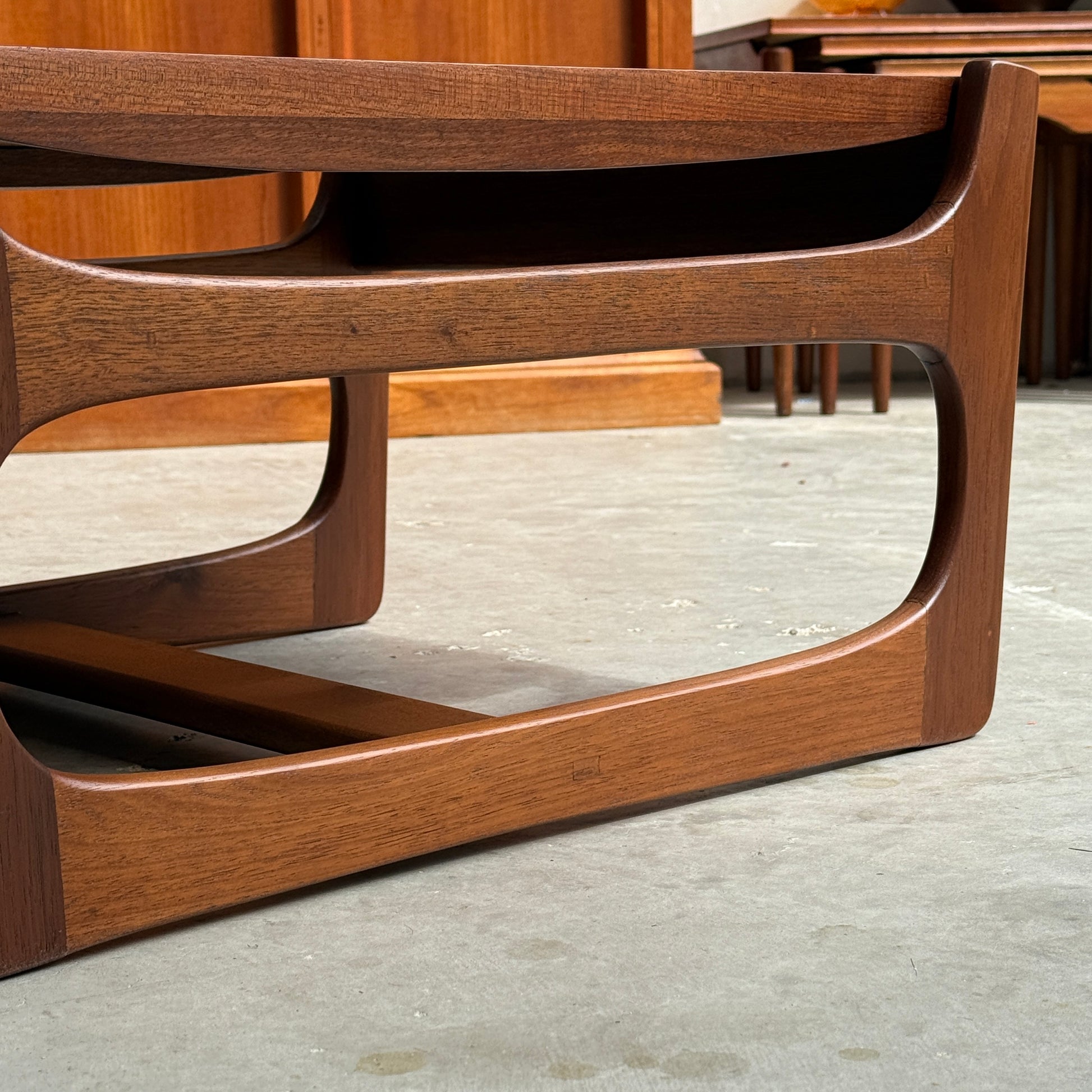 Close-up of an ultra rare Gerald Easden coffee table in Blackwood with a sleigh leg base (Rediscover), mid-century modern style, featuring curved edges and smooth finish on a concrete floor; another wooden piece is partially visible in the background.