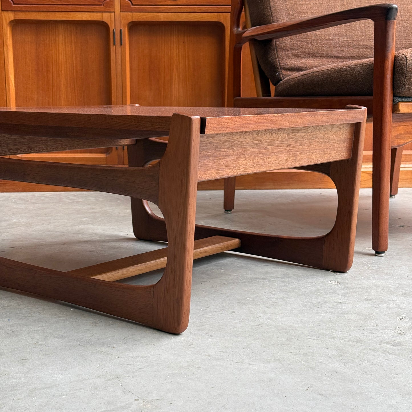 A close-up of the ultra rare Gerald Easden Coffee Table in Blackwood with a sleigh leg base, paired with a brown armchair on a concrete floor and wooden cabinets, captures the timeless elegance of Australian vintage style.