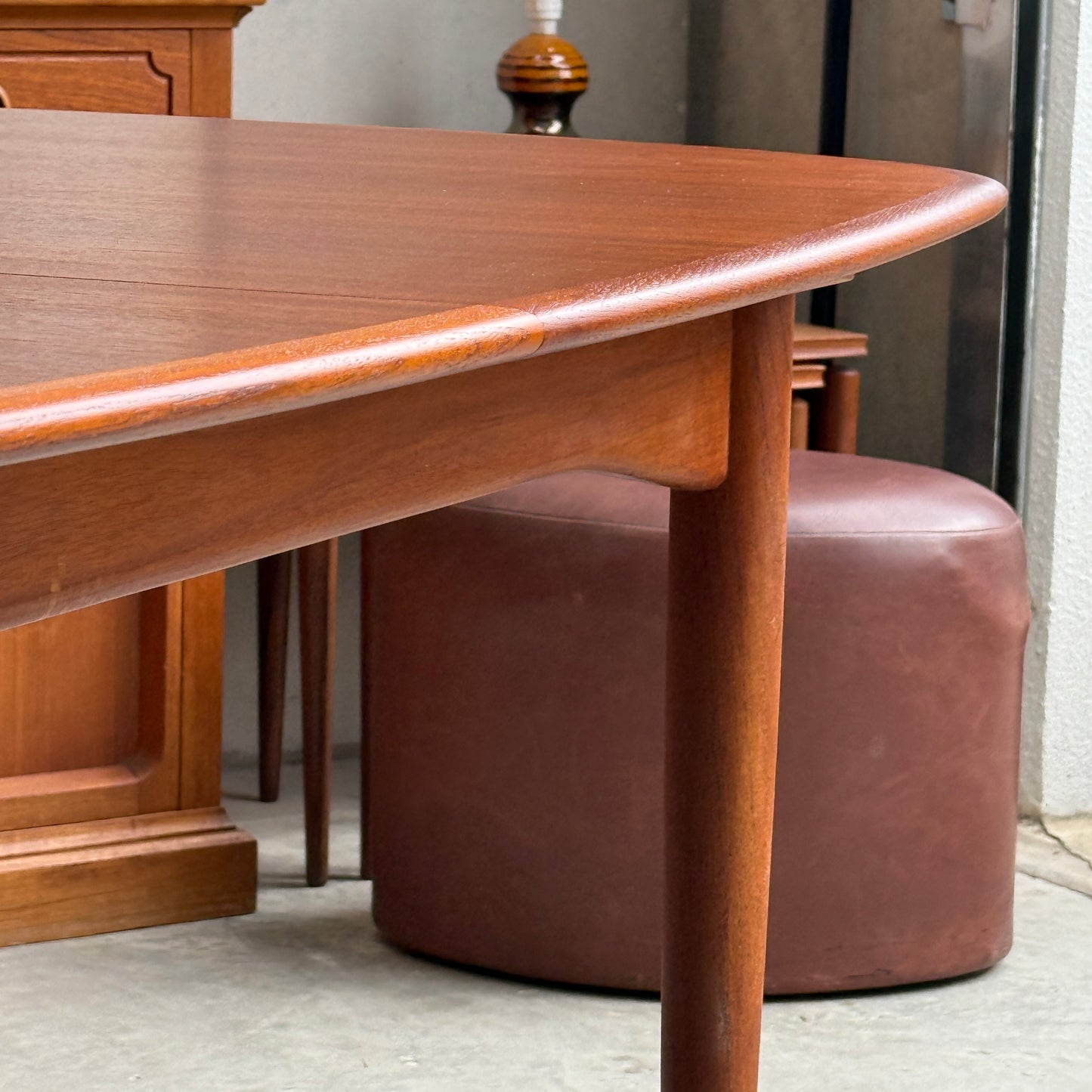 A close-up of the Fully Restored Parker No.92 D End Dining Table (Rediscover) with rounded edges stands on a concrete floor, with a brown ottoman, lamp, and wooden cabinet in the background for a mid-century modern touch.