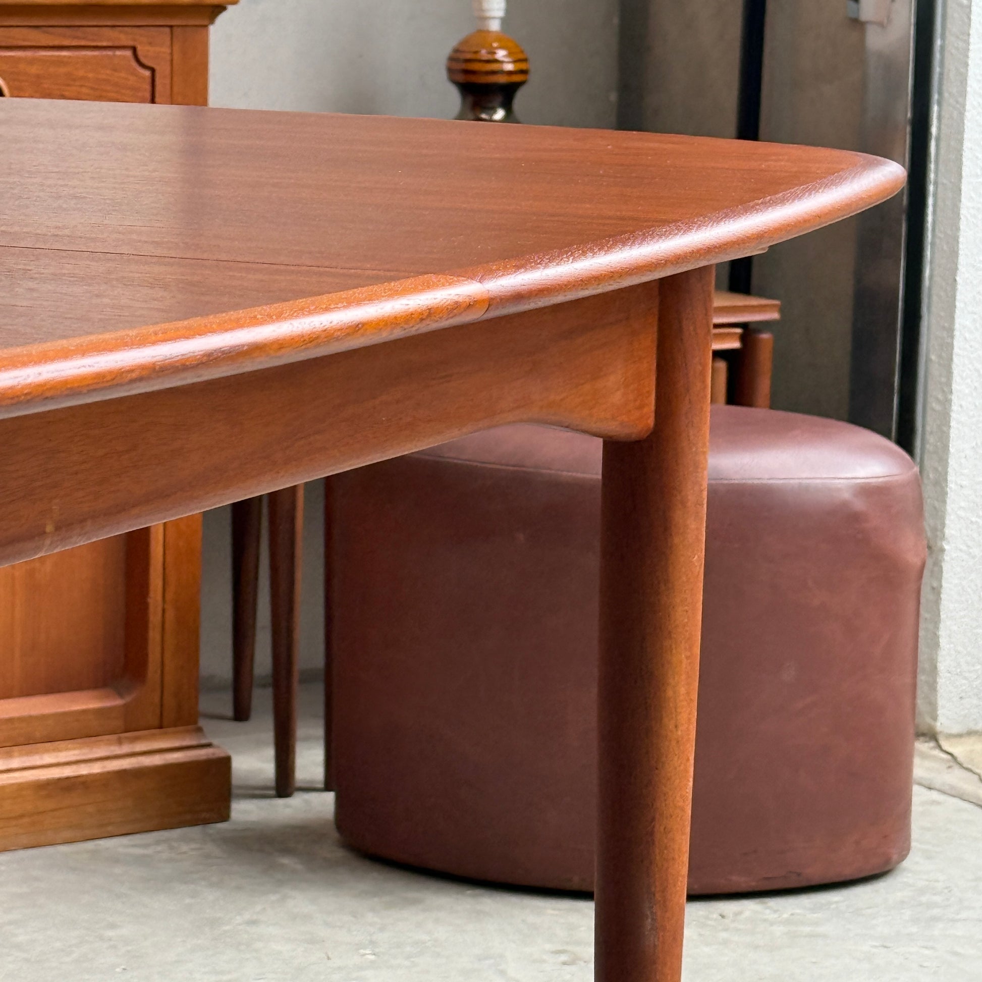 A close-up of the Fully Restored Parker No.92 D End Dining Table (Rediscover) with rounded edges stands on a concrete floor, with a brown ottoman, lamp, and wooden cabinet in the background for a mid-century modern touch.