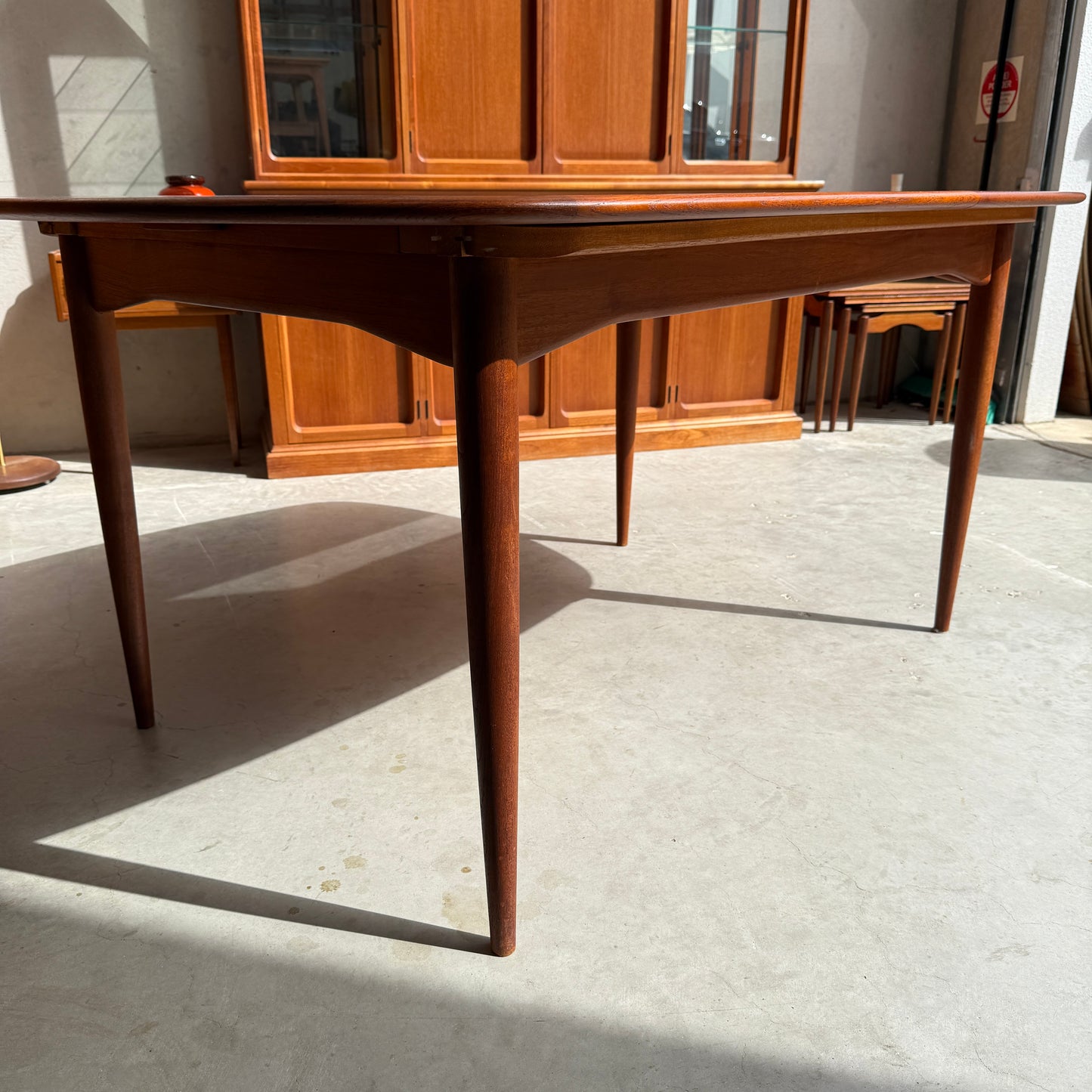 A Fully Restored Parker No.92 D End Dining Table (Rediscover) with tapered legs stands on a concrete floor in a sunlit room, while a wooden cabinet and nesting tables highlight vintage Australian furniture style in the background.