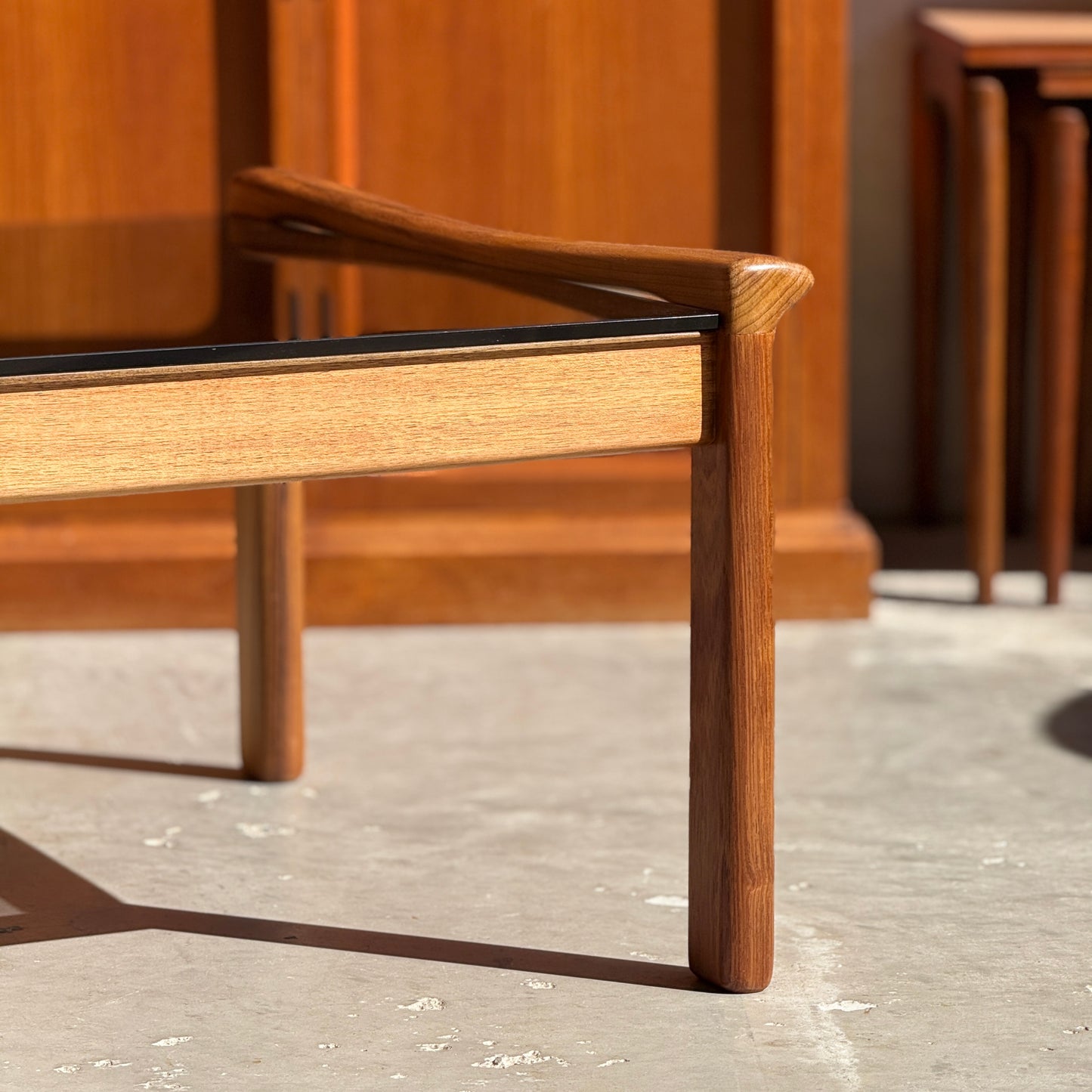 Close-up of a wooden chair’s armrest and leg with a restored frame in smooth mid-century style, sunlight casting shadows on the concrete floor; the Tessa TC 21 Coffee Table Frame Restored - (Revive) is visible in the background.