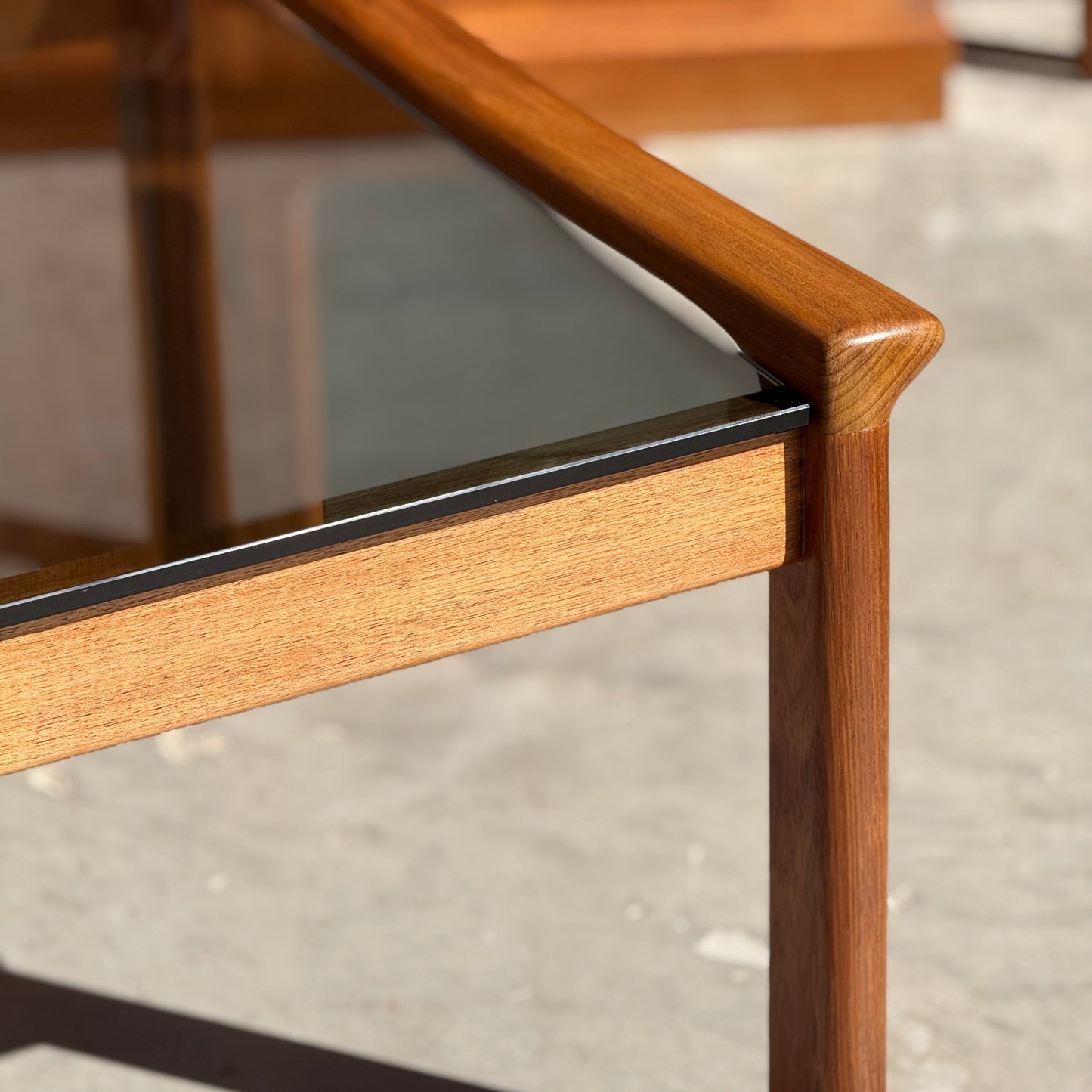 Close-up of a Tessa TC 21 Coffee Table Frame Restored - (Revive) with a glass top, smooth rounded edges, and warm brown wood grain, set on a concrete floor—ideal for fans of modern minimalist design.