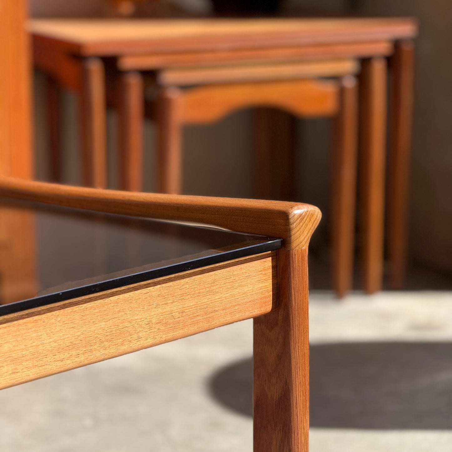Close-up of the Tessa TC 21 Coffee Table Frame Restored - (Revive) with glass top; a set of nested tables is blurred in the background, all basking in warm sunlight and highlighting modern minimalist design.