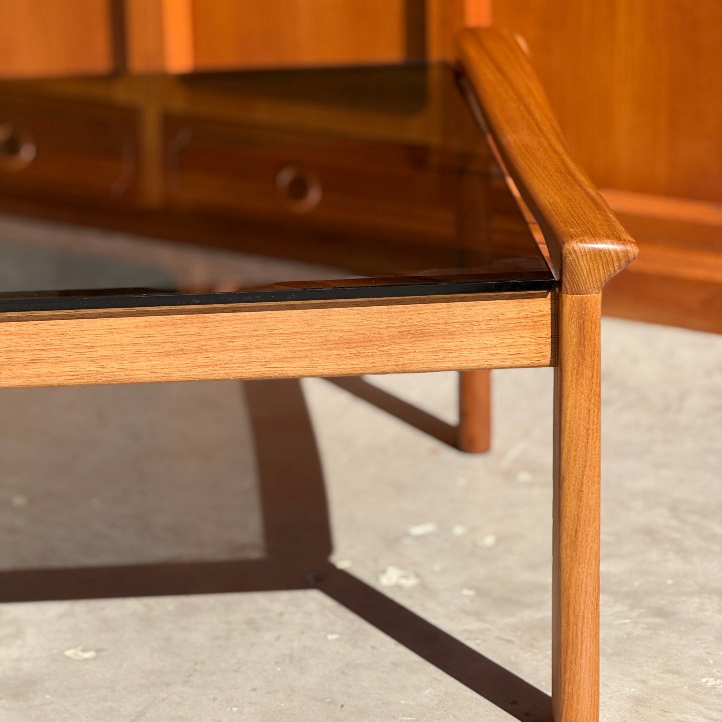 A close-up of the Tessa TC 21 Coffee Table Frame Restored - (Revive) with a glass top, highlighting its restored wood and angled legs. Sunlight enhances its modern minimalist look, with a blurred wooden cabinet in the background.