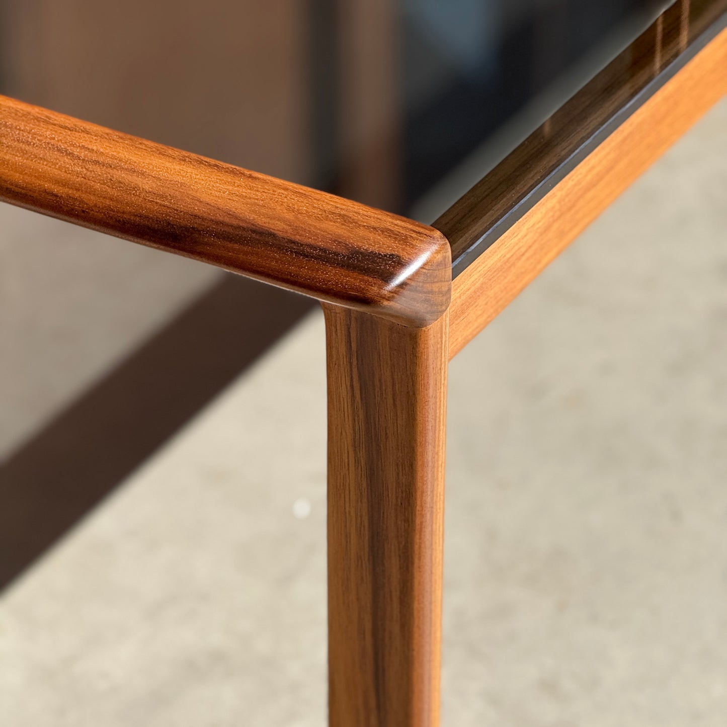 Close-up of the Tessa TC 21 Coffee Table Frame Restored - (Revive), featuring smooth, polished edges and a glass tabletop. Sunlight highlights the restored wood frame and minimalist design against a neutral background.