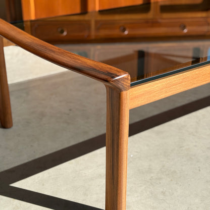 Close-up of the restored Tessa TC 21 Coffee Table Frame (Revive) with a glass top, showcasing smooth curved woodwork and modern minimalist design. Sunlight casts shadows on a concrete floor; a wooden drawer cabinet is blurred in the background.
