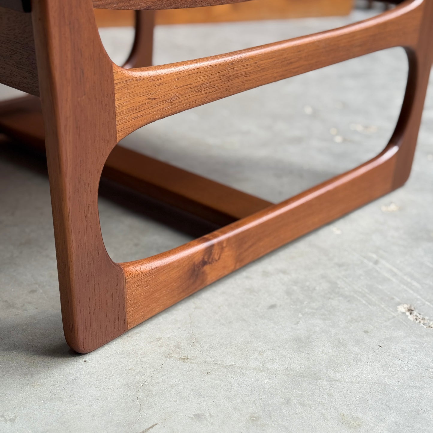 Close-up of a wooden leg with a curved, mid-century modern design from the Ultra Rare Gerald Easden Coffee Table in Blackwood Sleigh leg base (Rediscover), resting on smooth concrete. The warm finish highlights its rich grain and craftsmanship.