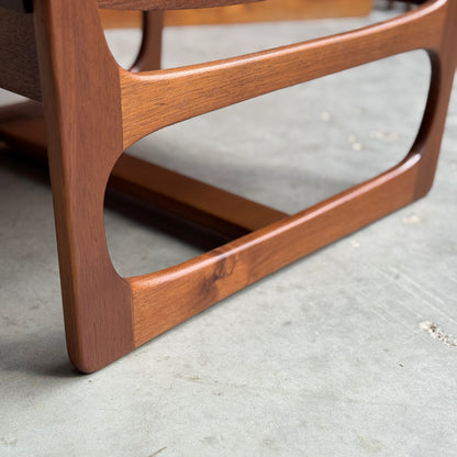 Close-up of a wooden leg with a curved, mid-century modern design from the Ultra Rare Gerald Easden Coffee Table in Blackwood Sleigh leg base (Rediscover), resting on smooth concrete. The warm finish highlights its rich grain and craftsmanship.