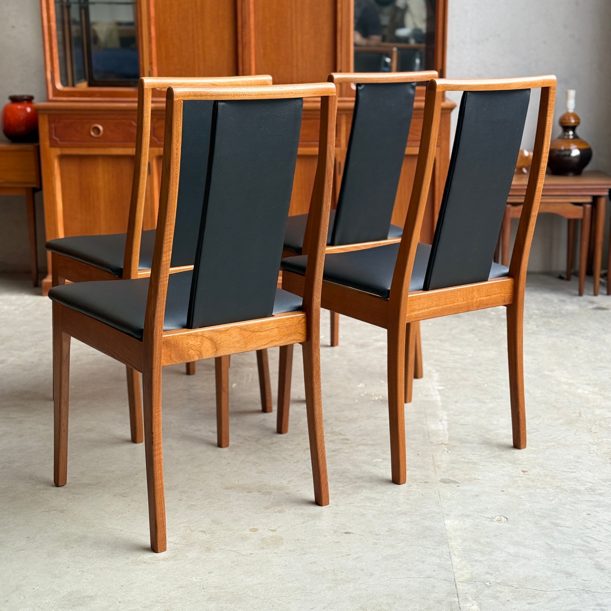 A set of 4 fully restored Noblett T-Back 'Boston' dining chairs in black vinyl is arranged together in a room with a wooden sideboard and mid-century modern decor.