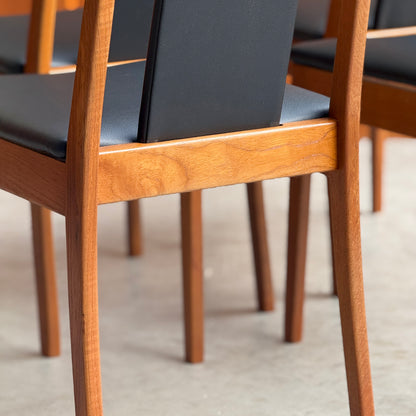 Close-up of NEW IN! Fully Restored Noblett T-Back 'Boston' Dining Chairs Set of 4 in black vinyl. The warm teak and padded seats highlight the natural grain and classic mid-century modern style, all arranged on a light-colored floor.