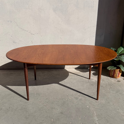 A fully restored Parker No.91 Fixed Top Oval Table (Rediscover) with slender legs stands on a concrete floor beside a plain light wall, sunlight casting shadows. A green leafy plant in a woven basket is partially visible to the right.