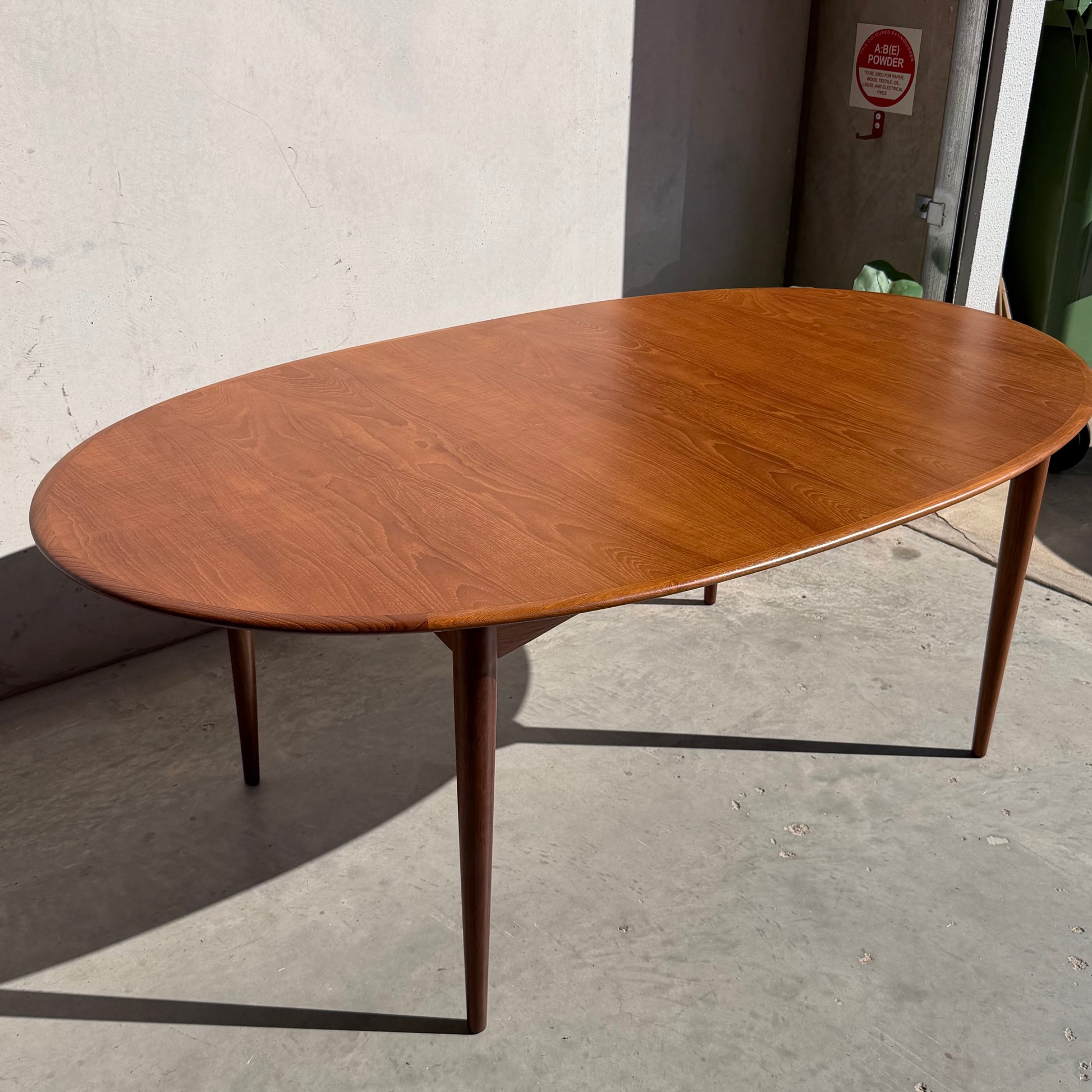 A fully restored Mid-Century Parker 91 Oval Teak Dining Suite with 6 Fler 64 Blackwood Chairs sits on a concrete floor near a light wall in a sunlit space, with a fire extinguisher sign visible in the background.