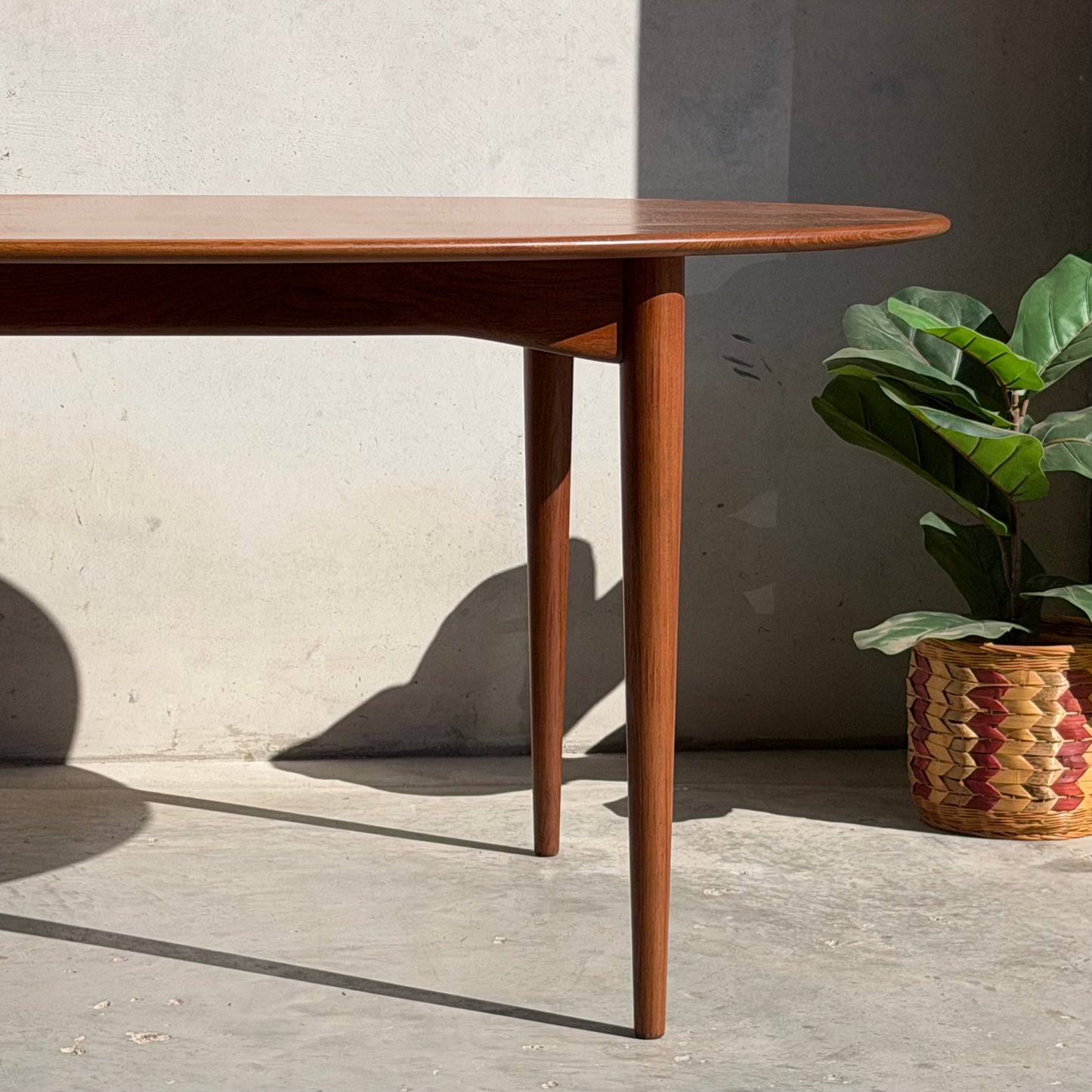 A Fully Restored Parker No.91 Fixed Top Oval Table (Rediscover) with rounded edges stands on a sunlit concrete floor beside a woven basket holding a green plant, set against a light-colored wall.