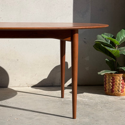 A fully restored mid-century Parker 91 oval teak dining suite with 6 Fler 64 blackwood chairs stands on a concrete floor near a potted green plant in a woven basket, sunlight casting gentle shadows along the wall behind.